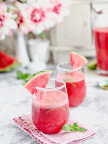 2 glasses of red drink sitting on a marble table garnished with watermelon wedges