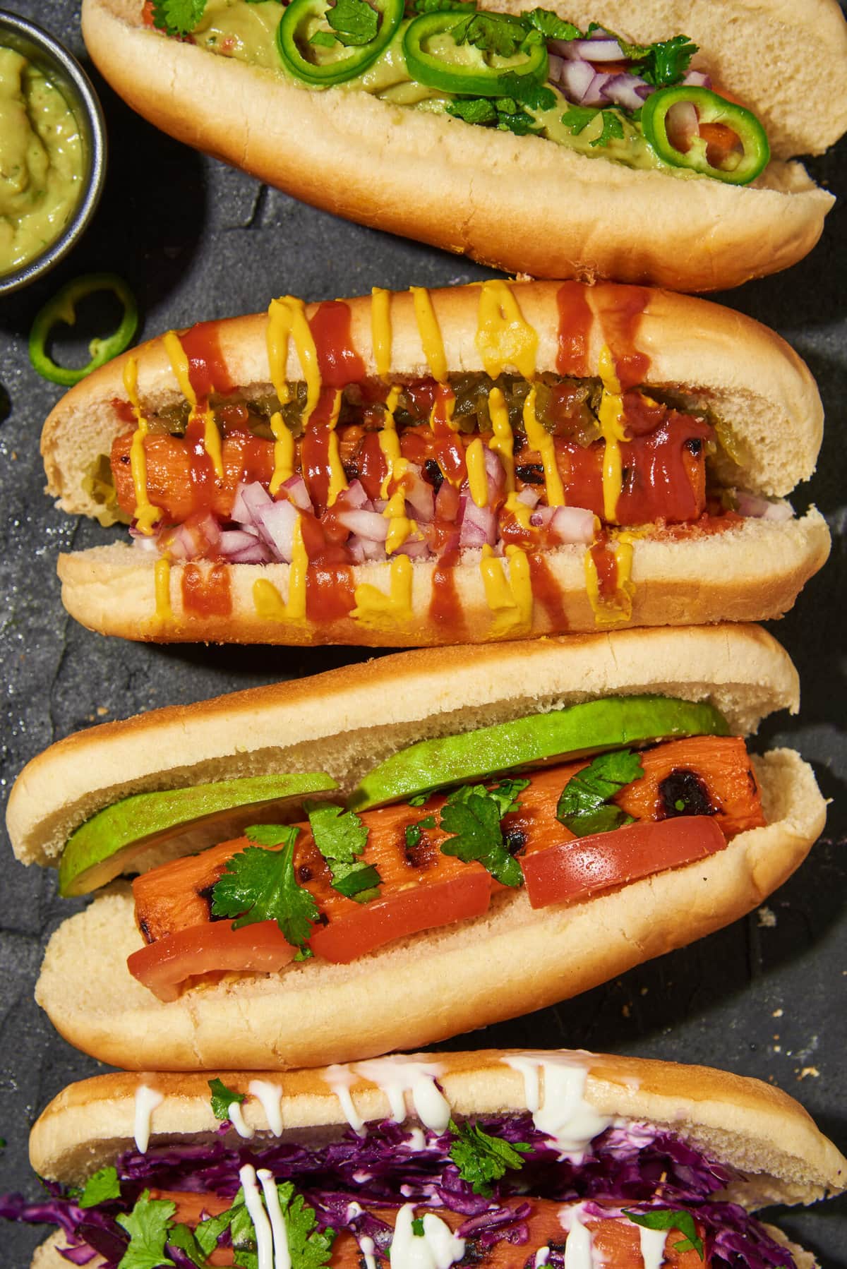 closeup overhead view of 4 grilled carrot dogs with different toppings on a dark table