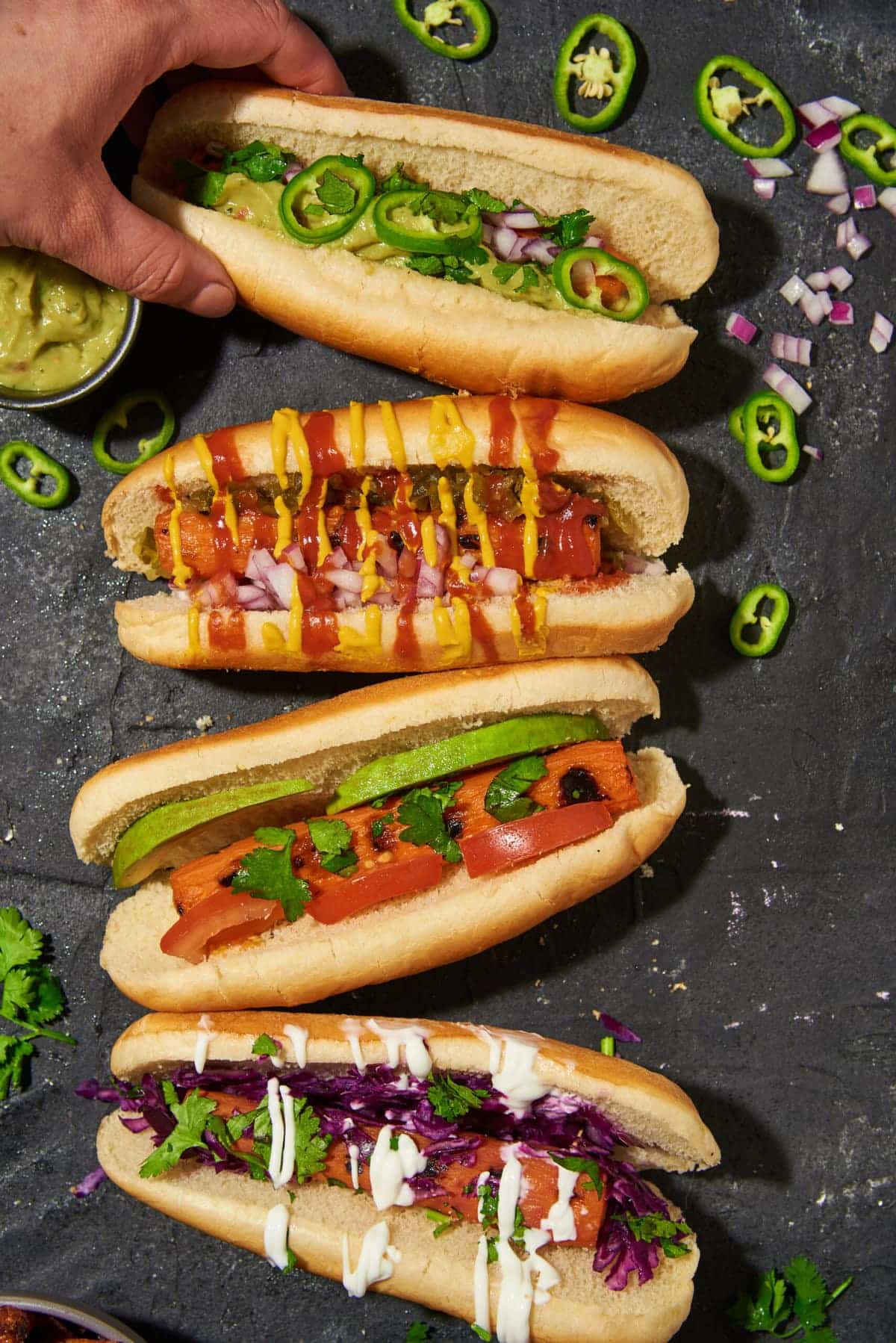 hand picking up one of four grilled carrot dogs on a dark table